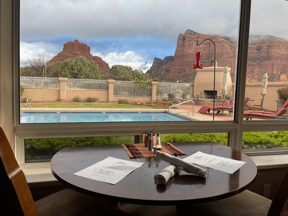 A table set for a meal overlooks a pool and dramatic red rock formations through a large window.