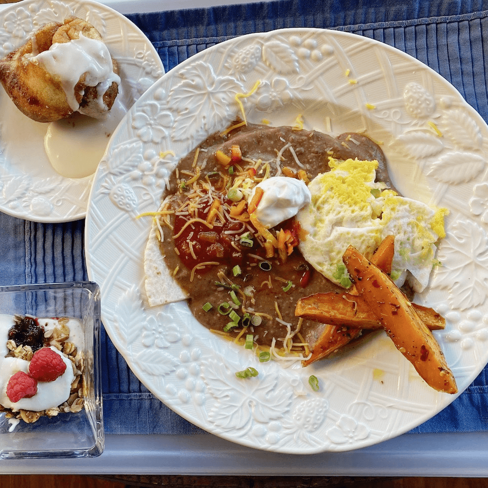 A plate of refried beans with cheese, salsa, eggs, and sweet potato fries, accompanied by a cinnamon roll and a bowl of yogurt with granola and berries.
